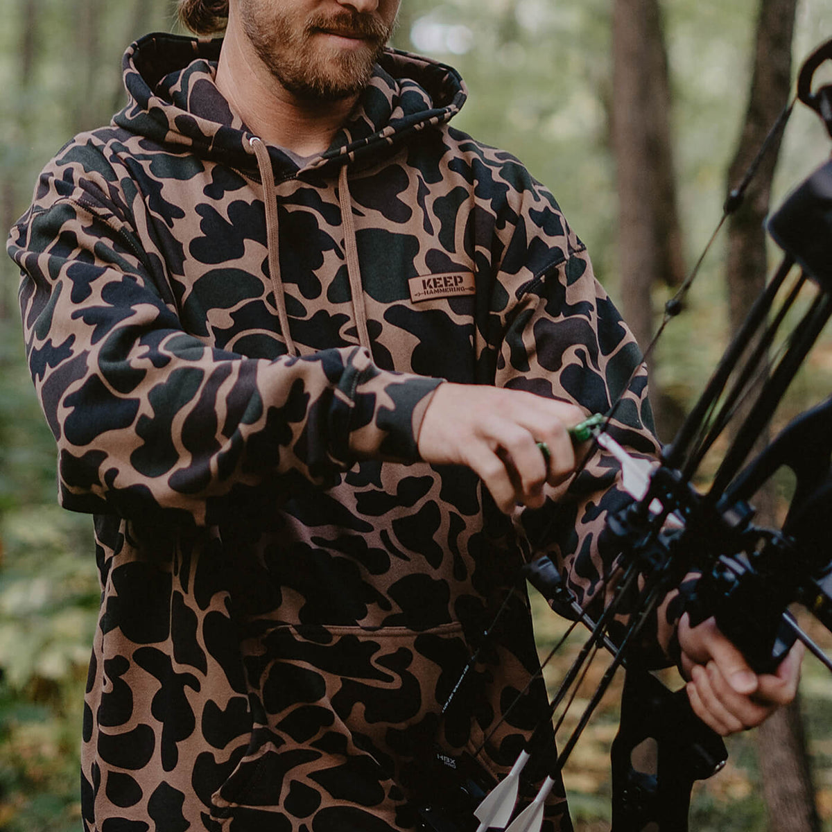 Keep Hammering OG Legacy Hoodie in duck camo, worn in the woods with a bow
